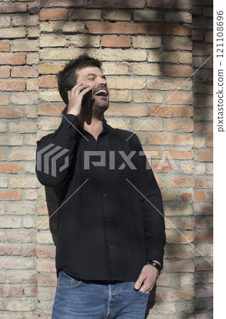 Man laughing while talking on the phone against a brick wall. 121108696