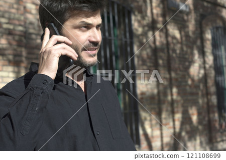 Man talking on the phone in sunlight near a brick building. 121108699
