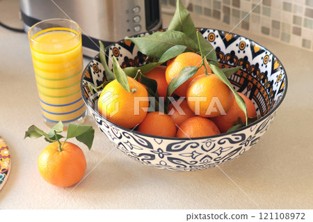 Clementines, mandarins with leaves in ceramic bowl. Fresh juice of citrus fruits in glass on table in kitchen. Mosaics and pattern on dishes.  121108972