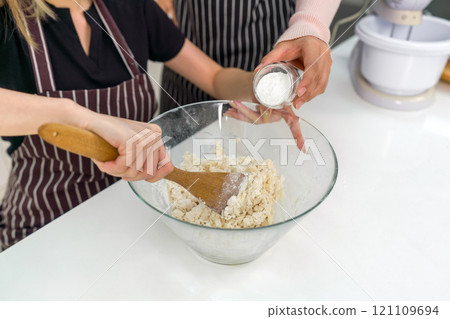 Two women make dessert in the kitchen. One people stirring a white cake batter or icing mixture. The other pouring baking powder in a clear glass bowl. 121109694