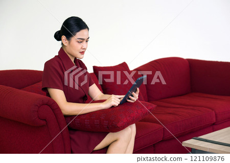 Young asian businesswoman in red dress working on red sofa at home. Tablet computer and cushion are on her lap. Young asian businesswoman in red dress working on red sofa at home. Tablet computer and cushion are on her lap. 121109876