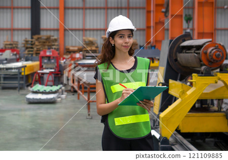 A woman wearing a safety vest and hardhat stand in a factory. She holds a clipboard and looks confident. A woman wearing a safety vest and hardhat stand in a factory. She holds a clipboard and looks confident. 121109885