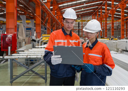 Two worker in safety gear look at a laptop computer, standing in a large factory. They are checking equipment and discussing plan. Two worker in safety gear look at a laptop computer, standing in a large factory. They are checking equipment and discussing plan. 121110293