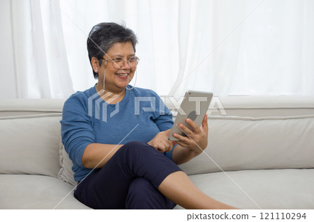 Asian gray hair old woman looking at tablet computer monitor, smiling while sit on sofa in the living room. Asian gray hair old woman looking at tablet computer monitor, smiling while sit on sofa in the living room. 121110294