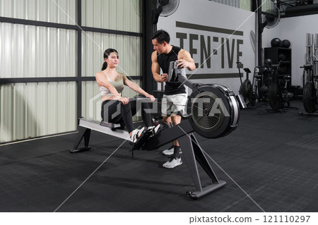An athletic woman in sportswear is engaged in an intense workout session on a rowing machine with a trainer inside a well-equipped gym. An athletic woman in sportswear is engaged in an intense workout session on a rowing machine with a trainer inside a well-equipped gym. 121110297