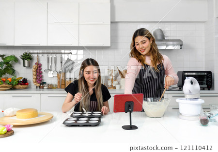 Two young women in an apron make dessert in the kitchen while looking at online recipe guide book on tablet computer. 121110412