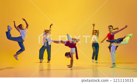 Dynamic image of stylish teen girls wearing casual attire, dancing hip hop, training against yellow background in neon light 121113015