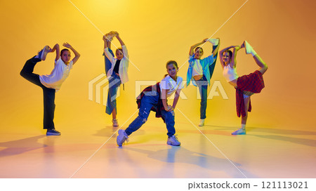 Flexibility and talent. Group of teen girls in casual stylish attire in motion, dancing hip hop together against yellow background in neon light 121113021