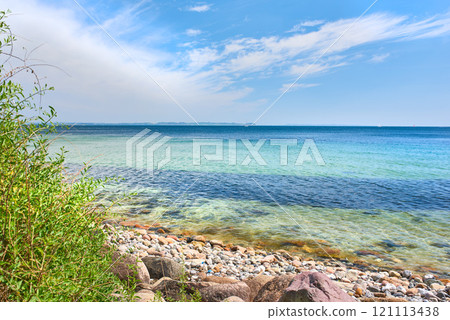 Copyspace at sea with a cloudy blue sky background. Calm ocean waves washing onto stones at an empty beach shore with sailboats cruising in the horizon. Scenic landscape for a relaxing summer holiday Copyspace at sea with a cloudy blue sky background. Calm ocean waves washing onto stones at an empty beach shore with sailboats cruising in the horizon. Scenic landscape for a relaxing summer holiday 121113438