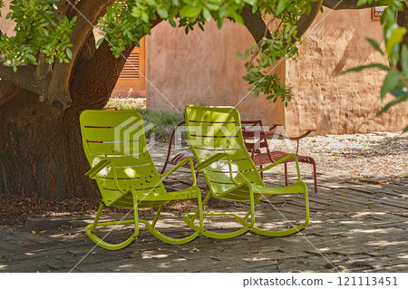 Two green vintage courtyard rocking chairs under a shady tree in a secluded and private garden at home. Patio furniture and seating set to enjoy and relax in summer. Lounging and feeling cosy outside Two green vintage courtyard rocking chairs under a shady tree in a secluded and private garden at home. Patio furniture and seating set to enjoy and relax in summer. Lounging and feeling cosy outside 121113451