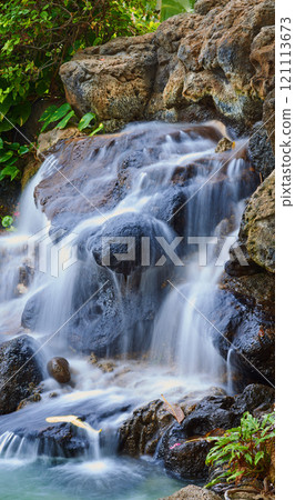 Serene pond with a waterfall over rocks. Landscape of a small natural spring near a clear pool on a mountain trail. Relaxing peaceful nature scene. Calm background of water flowing on stone outside 121113673