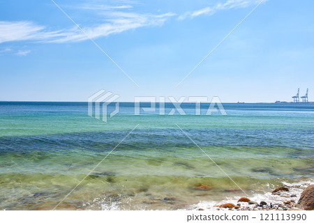 Landscape view of a blue ocean with clear skies on the horizon on a warm summer day. Little waves crash on a quiet calm rocky shore in the mediterranean sea. Panoramic coastline location for tourist 121113990