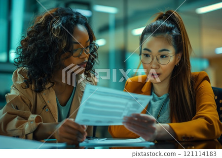 Two women engaged in a discussion over documents in a modern office setting. 121114183