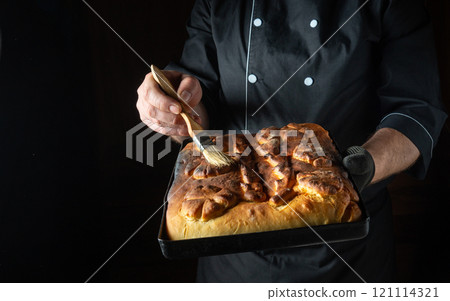 A skilled chef applies a delicate glaze to a freshly baked loaf of bread while holding it in a dark kitchen. The aroma of warm yeast fills the air as the crust shines A skilled chef applies a delicate glaze to a freshly baked loaf of bread while holding it in a dark kitchen. The aroma of warm yeast fills the air as the crust shines 121114321
