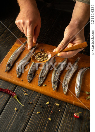 Skilled hands delicately arrange fresh fish on a wooden board, expertly adding spices from a small spoon while vibrant chilies and herbs enhance the culinary atmosphere. 121114323