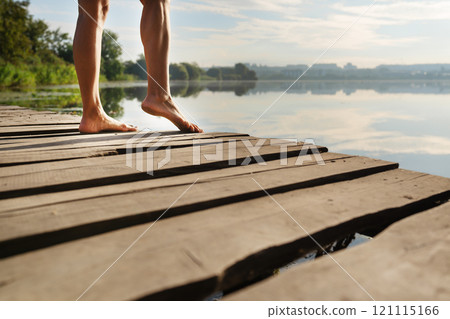 Close-up of bare feet on a wooden dock with a serene lake and trees in the background under a bright sky Close-up of bare feet on a wooden dock with a serene lake and trees in the background under a bright sky 121115166