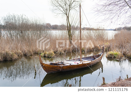 A wooden Viking-style boat resting on a still lake surrounded by reeds and bare trees under a cloudy sky A wooden Viking-style boat resting on a still lake surrounded by reeds and bare trees under a cloudy sky 121115176