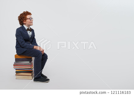 Smiling redhead child schoolboy sitting on stack of books and looking up against white studio wall  121116183
