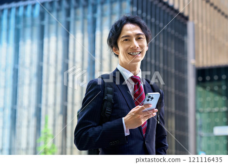 A businessman in a suit carrying a backpack operating a smartphone A businessman in a suit carrying a backpack operating a smartphone 121116435