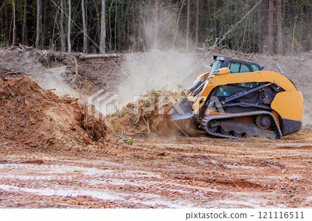 Heavy bulldozer machinery operates to clear section of land, creating dust clouds as it churns through dirt in construction site near at forested area 121116511