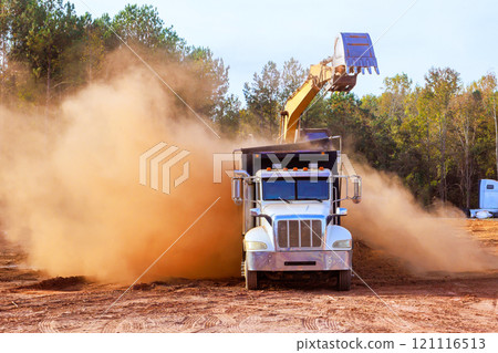 Construction truck is unloading dirt while large excavator scoops material dust clouds billow indicating active earthmoving operations Construction truck is unloading dirt while large excavator scoops material dust clouds billow indicating active earthmoving operations 121116513