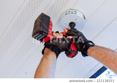 Using cordless drill with hole saw attachment, gloved hand cuts circular hole in white ceiling to install new light fixture Using cordless drill with hole saw attachment, gloved hand cuts circular hole in white ceiling to install new light fixture 121116551