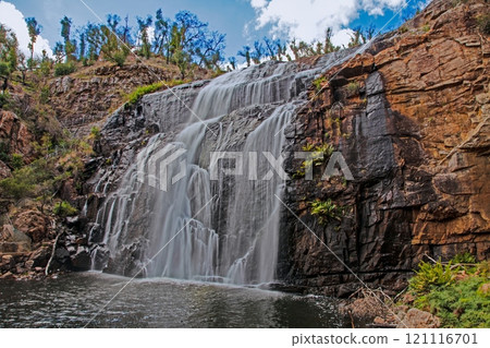 Waterfall cascading down rugged cliffs surrounded by lush greenery under a bright blue sky 121116701