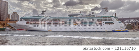 Large cruise ship docked at the harbor with dramatic cloudy sky and urban buildings in the background 121116710
