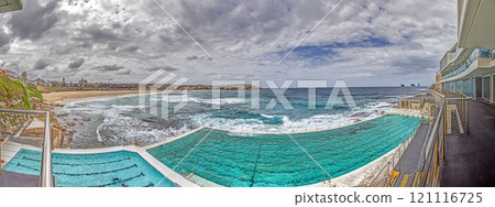 Panoramic view of Bondi Beach in Sydney, Australia, featuring the iconic Bondi Icebergs pool and ocean waves under a cloudy sky Panoramic view of Bondi Beach in Sydney, Australia, featuring the iconic Bondi Icebergs pool and ocean waves under a cloudy sky 121116725