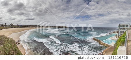 Panoramic view of Bondi Beach in Sydney, Australia, featuring the iconic Bondi Icebergs pool and ocean waves under a cloudy sky Panoramic view of Bondi Beach in Sydney, Australia, featuring the iconic Bondi Icebergs pool and ocean waves under a cloudy sky 121116726