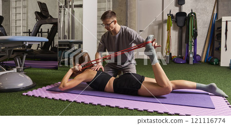 Woman lying on mat, stretching with red resistance band, guided by trainer during rehabilitation workout in gym. 121116774