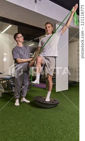 Young man using resistance band while balancing on trainer disk, assisted by professional during rehabilitation session. 121116775