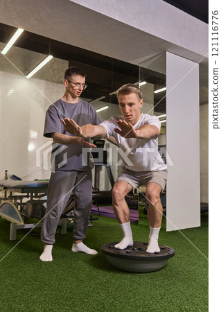 Man performing squats on balance trainer, receiving support and feedback from trainer during rehabilitation session. 121116776
