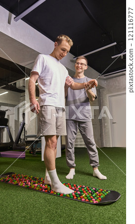 Man walking barefoot on colorful reflexology massage board, assisted by trainer for therapeutic foot stimulation. 121116777