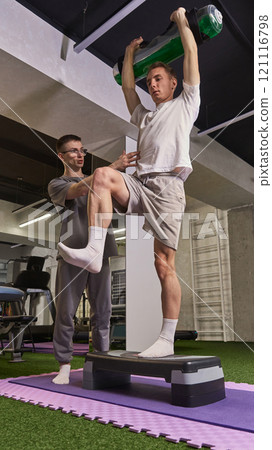 Man performing step-up exercise while holding water-filled weight, supervised by trainer for rehabilitation and balance improvement. Man performing step-up exercise while holding water-filled weight, supervised by trainer for rehabilitation and balance improvement. 121116798