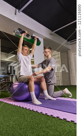 Man lifting water-filled weight overhead while seated on balance ball, guided by professional trainer for rehabilitation. Man lifting water-filled weight overhead while seated on balance ball, guided by professional trainer for rehabilitation. 121116800