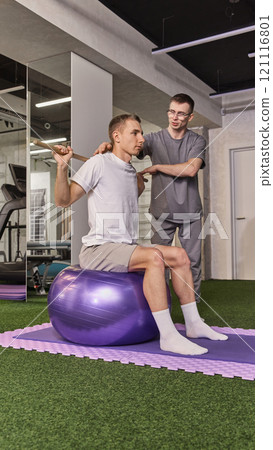 Man performing back exercise on balance ball with wooden stick, carefully supported by trainer in recovery session. Man performing back exercise on balance ball with wooden stick, carefully supported by trainer in recovery session. 121116801