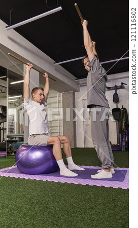 Man lifting wooden stick overhead while balancing on exercise ball, assisted by trainer in rehabilitation-focused gym. Man lifting wooden stick overhead while balancing on exercise ball, assisted by trainer in rehabilitation-focused gym. 121116802
