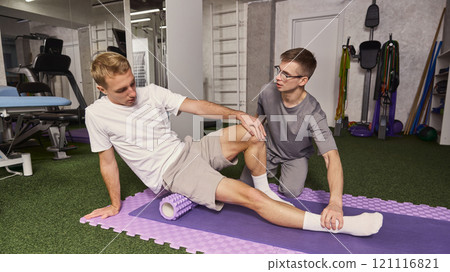 Physiotherapist assisting patient in using foam roller for leg recovery during rehabilitation session in gym. 121116821
