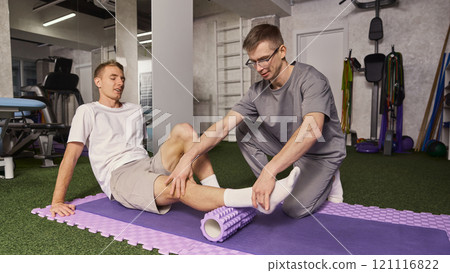 Young man, patient sitting on foam roller with physiotherapist guiding the movement for muscle recovery and rehabilitation exercises. 121116822
