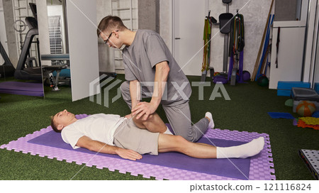 Man, physiotherapist stretching young man, patients leg, performing leg mobility while lying on mat in gym environment Man, physiotherapist stretching young man, patients leg, performing leg mobility while lying on mat in gym environment 121116824