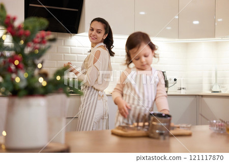 Cute little child girl in apron helping mom bake Christmas cookies in the kitchen, focus on mom 121117870