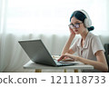 A woman wearing headphones and glasses is typing on a laptop 121118733