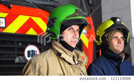Portrait of male firefighters in helmets and protective uniforms standing near big red car at department. Young confident firemen in full equipment posing against background of fire engine at station Portrait of male firefighters in helmets and protective uniforms standing near big red car at department. Young confident firemen in full equipment posing against background of fire engine at station 121119006