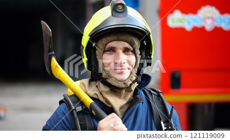 Portrait of happy male fireguard with axe standing near a fire engine. Young smiling fireman looking at camera in full equipment after fire near big red truck. Saving lives and heroic profession 121119009