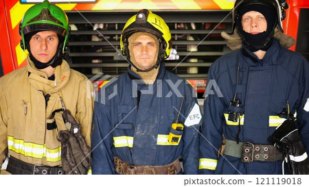 Portrait of male firefighters in helmets and protective uniforms standing near a big red car. Young confident firemen in full equipment posing against the background of a fire engine at station 121119018