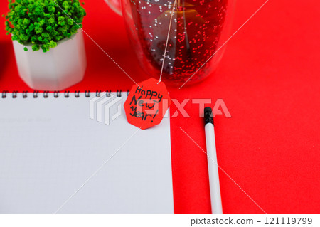 A white notebook with a black spring on which, a white pencil lies nearby, on top there is a green plant in a pot and a cup of tea with a red label of a happy New Year and on a red background close-up A white notebook with a black spring on which, a white pencil lies nearby, on top there is a green plant in a pot and a cup of tea with a red label of a happy New Year and on a red background close-up 121119799