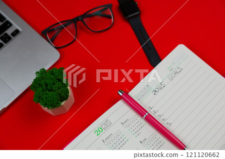 A close-up on a red background with a slope lies a pink diary notebook in which it is written: Goals for 2025, inside it is a calendar for 2025 and there is a pink pen, an open laptop, part of the A close-up on a red background with a slope lies a pink diary notebook in which it is written: Goals for 2025, inside it is a calendar for 2025 and there is a pink pen, an open laptop, part of the 121120662