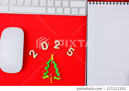 In the center of the photo on a red background there are wooden numbers 2025 and a Christmas tree made of office nails, on the side there is a white notebook with a black spring at the top and a 121121102