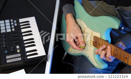 Male musician skillfully strums a mint electric guitar while a MIDI keyboard controller sits nearby during a songwriting session in his home studio Male musician skillfully strums a mint electric guitar while a MIDI keyboard controller sits nearby during a songwriting session in his home studio 121121324